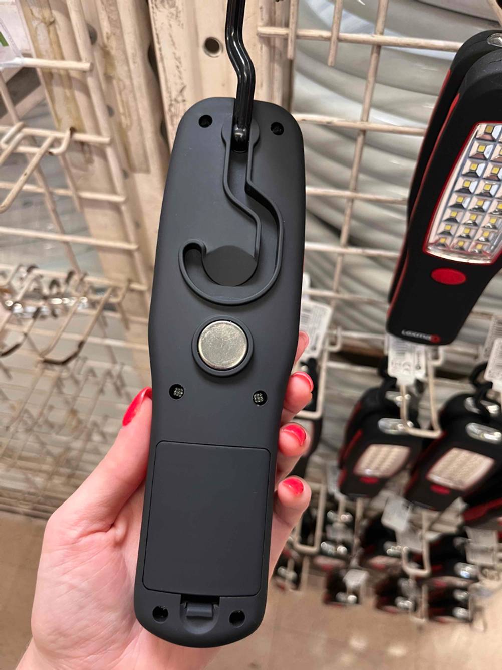 LED-Strahler im Test: Hand hält schwarzen LED-Strahler vor Kaufregal. Blick auf die Rückseite mit Magnet und Haken-Aufhängung sowie Batteriefach.