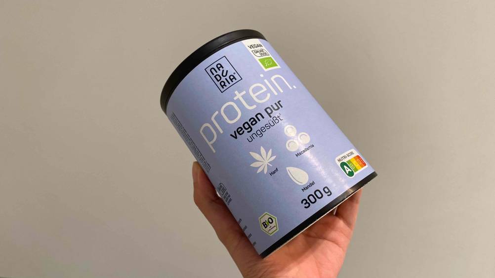Bio-Proteinpulver im Test: Eine Hand hält eine Dose Pulver vor einer hellen Wand hoch