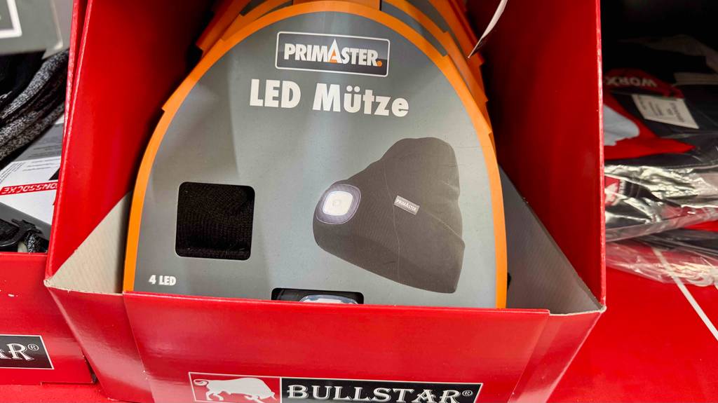 Die getestete LED-Mütze in Schwarz innerhalb eines grau-orangefarbenen Verpackungskartons befindet sich auf einem Verkaufsregal.