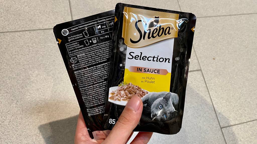 Sheba-Katzenfutter getestet: Zwei Packungen Katzenfutter in der Hand -Sorte: Huhn mit Pulet in Sauce.