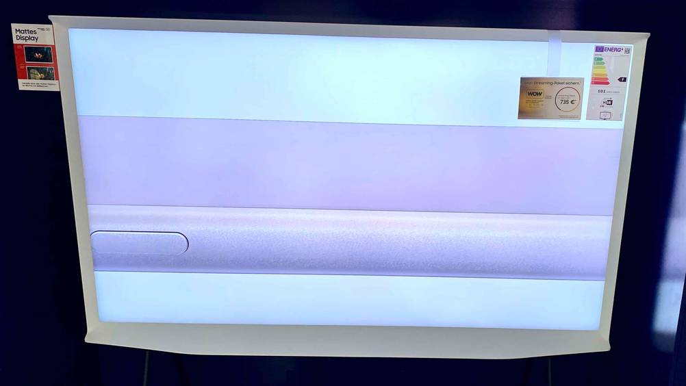 Ein angeschalteter 65-Zoll-Fernseher von vorne mit weißem Rahmen und Infoaufklebern.