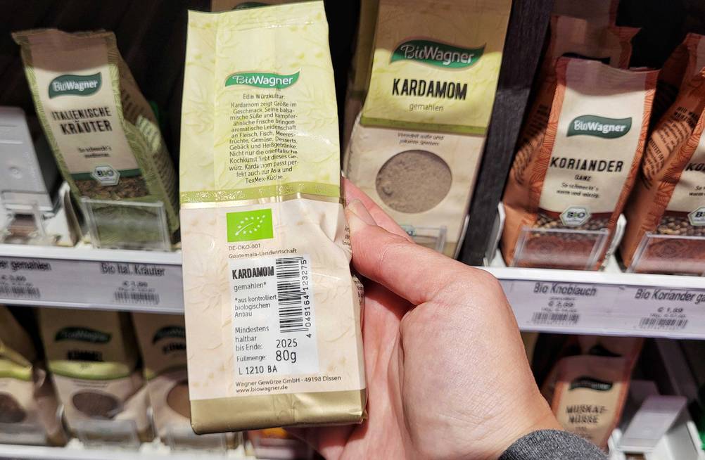 Kardamom im Test: eine Packung Kardamom wird mit der Rückseite ersichtlich vor ein Regal gehalten.