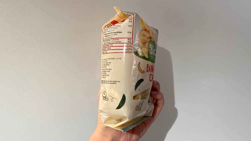 Eine Hand hält eine Packung getesteter Bananenchips seitlich vor eine Wand.