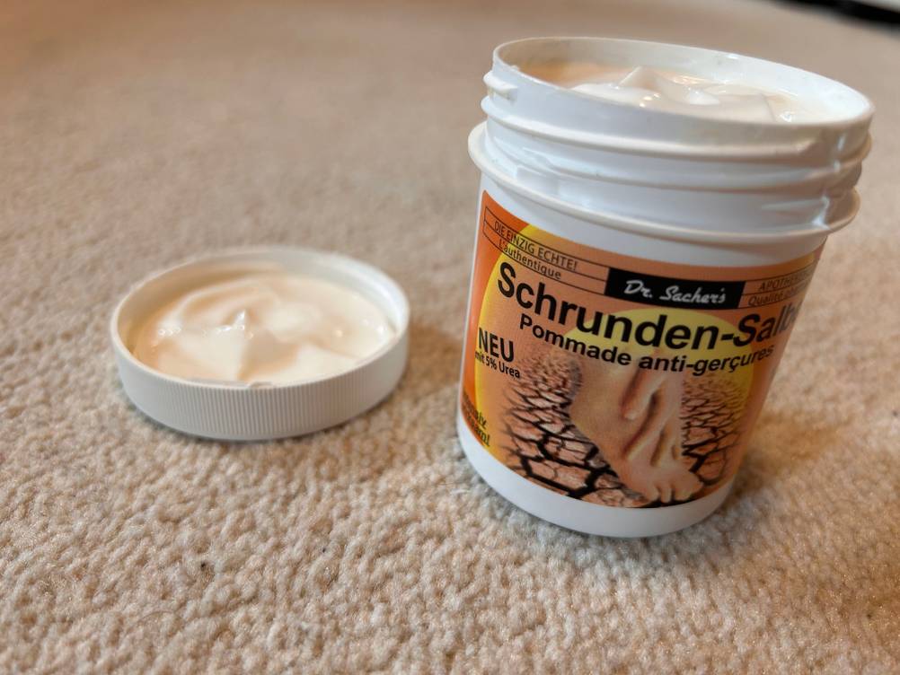 Auf einem Tepppichboden steht eine geöffnete Dose der getesteten Schrundensalbe, ihr Deckel liegt daneben.