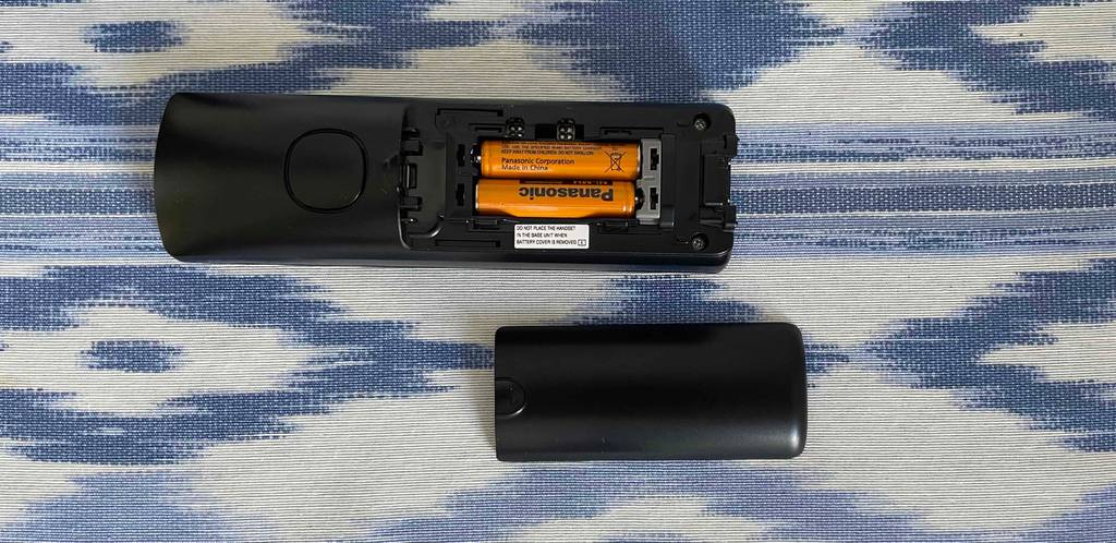 Ein Telefon mit offenem Batteriefach liegt neben dem Batteriefach-Deckel auf blau-weiß-meliertem Stoff.