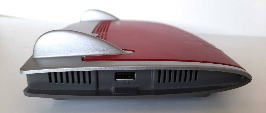 Fritzbox-Test: Seitenansicht von einem Modell in Rot, Silber und Schwarz mit Fokus auf die integrierte USB-Buchse vor weißem Hintergrund.