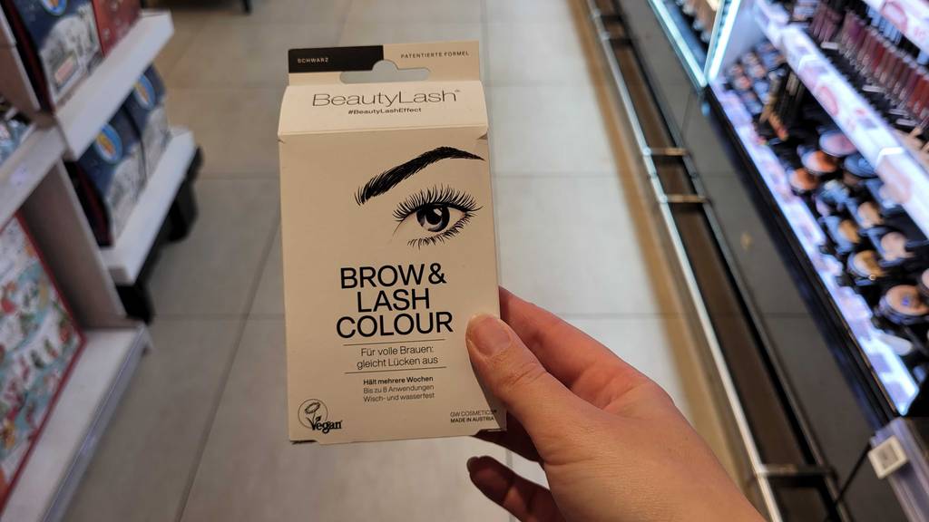 Eine getestete Wimpernfarbe in der Farbe Schwarz wird in der Hand vor einem Regal gehalten.