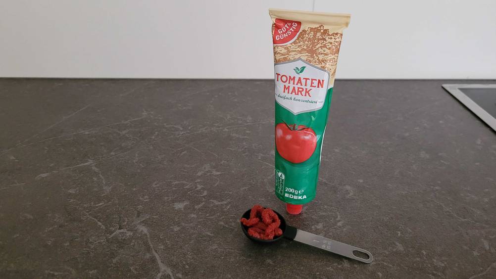 Im Test: Auf einer dunklen Arbeitsplatte steht eine Tube Tomatenmark , davon ein gefüllter Löffel.