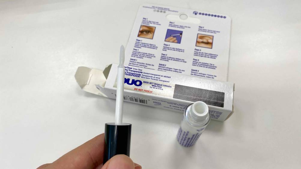Wimpernkleber im Test. Verpackung von Wimpernkleber liegend und davor wird der Applikator des geöffneten Wimpernklebers gehalten.