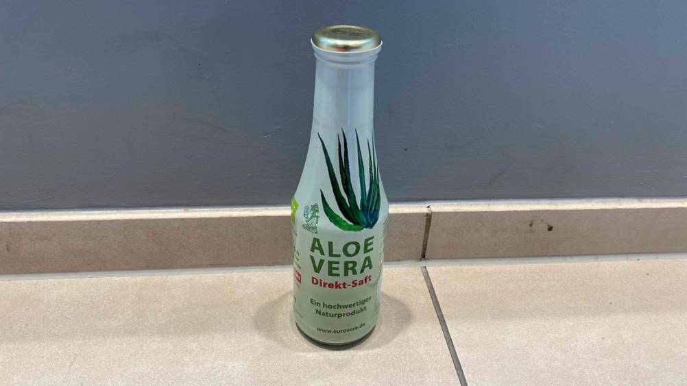 Aloe-vera-Saft getestet: Vor einer blauen Wand auf einem hellen Untergrund steht eine Flasche.
