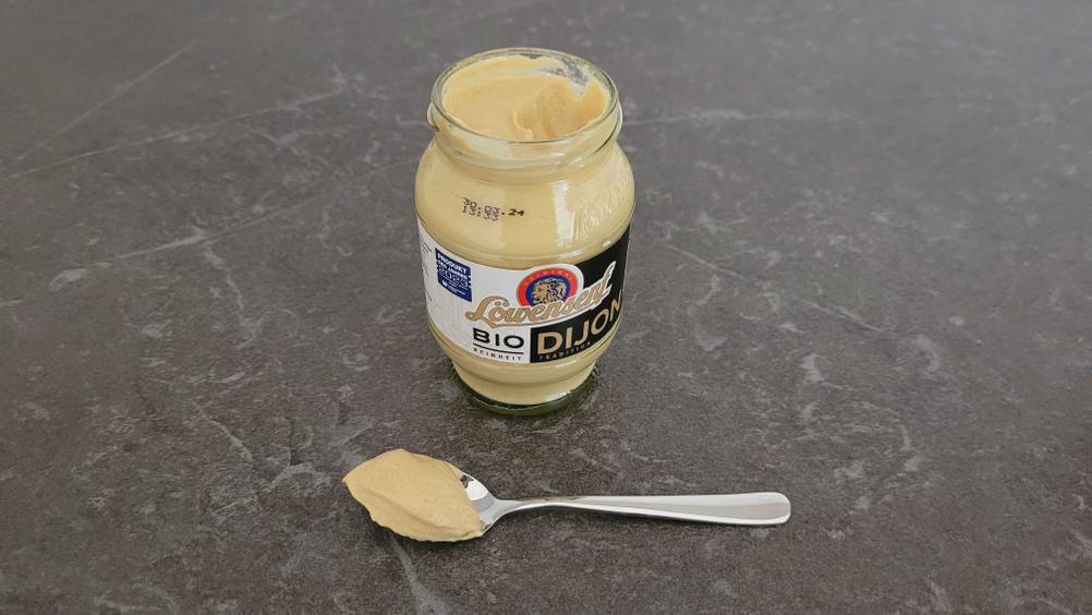 Dijon Senf im Test: Ein Löffel mit Senf liegt vor einem geöffneten Glas Senf auf einer dunklen Oberfläche