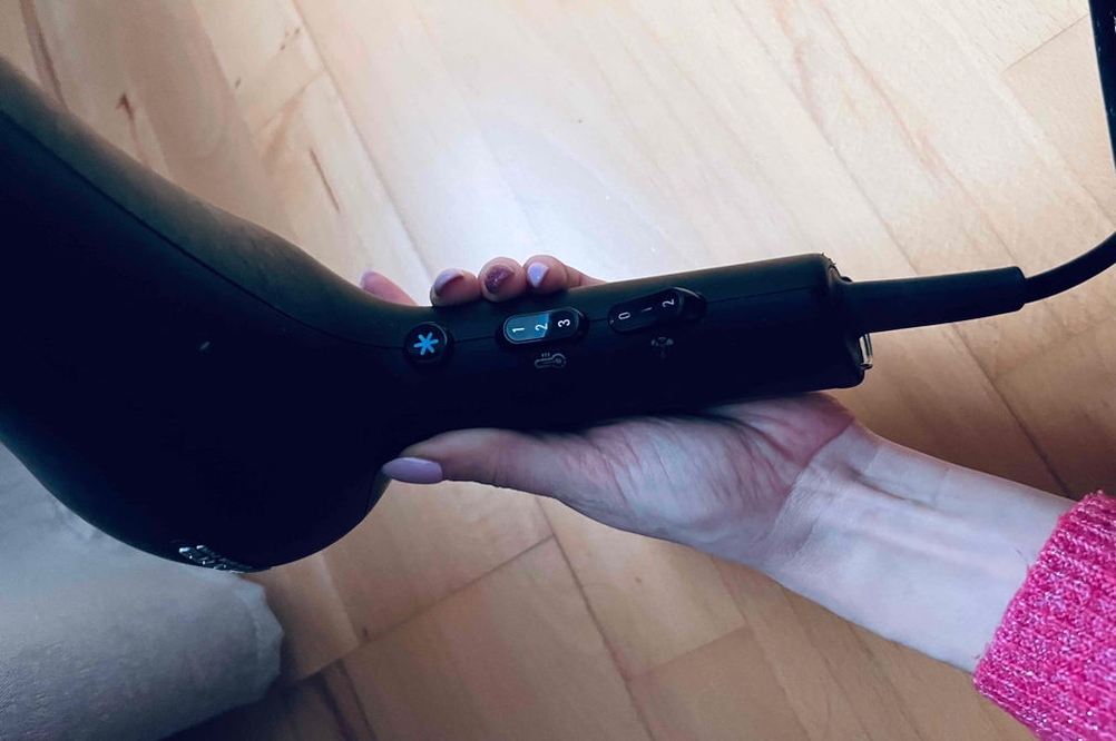  In einer Hand befindet sich ein Ionen-Haartrockner vor einer Holzoberfläche.