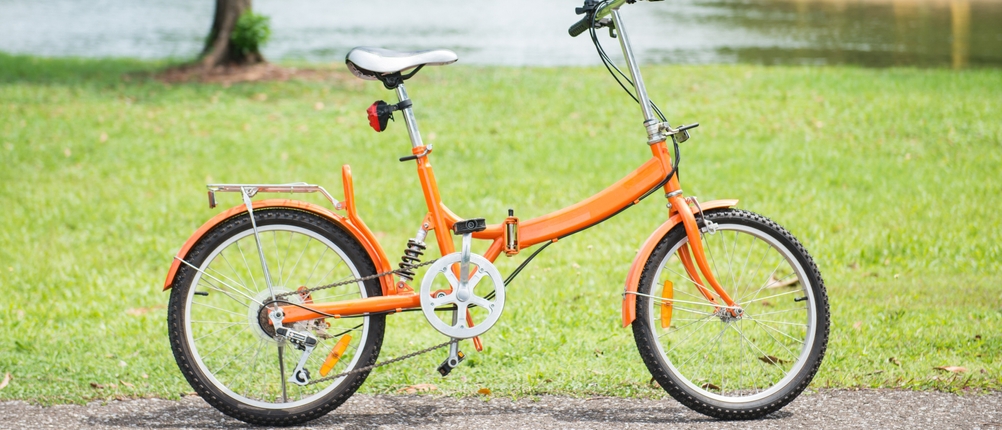 Ein orangenes Klapprad steht in einem Park am Rand eines Weges.