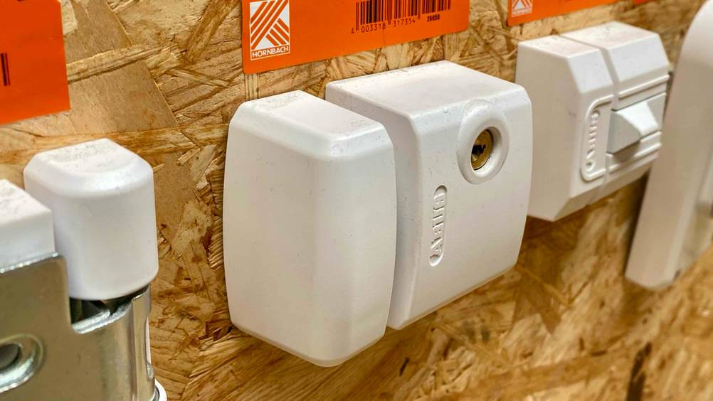 Eine getestete Fenstersicherung in Nahaufnahme an einem Holzbrett zwischen anderen Produkten befestigt.