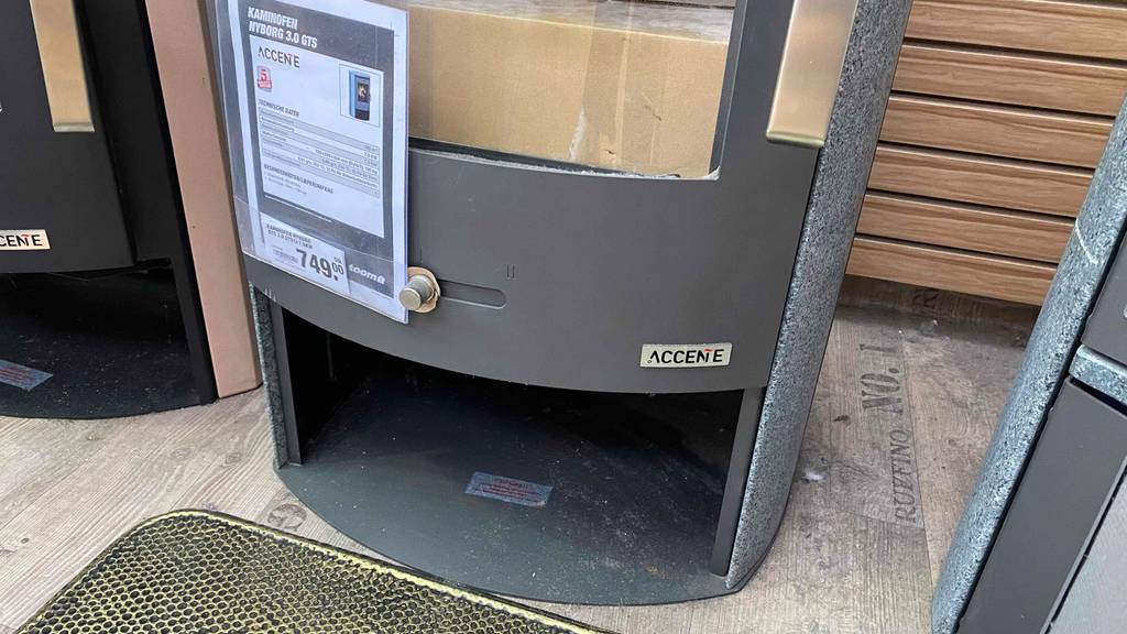 Detailansicht eines Accente Kaminofens im Test: mit Fokus auf das unten befindliche Holzfach des anthrazit-grauen Zeitbrandofens, mit Linksanschlag und Glasfenster, inklusive Preisschild.