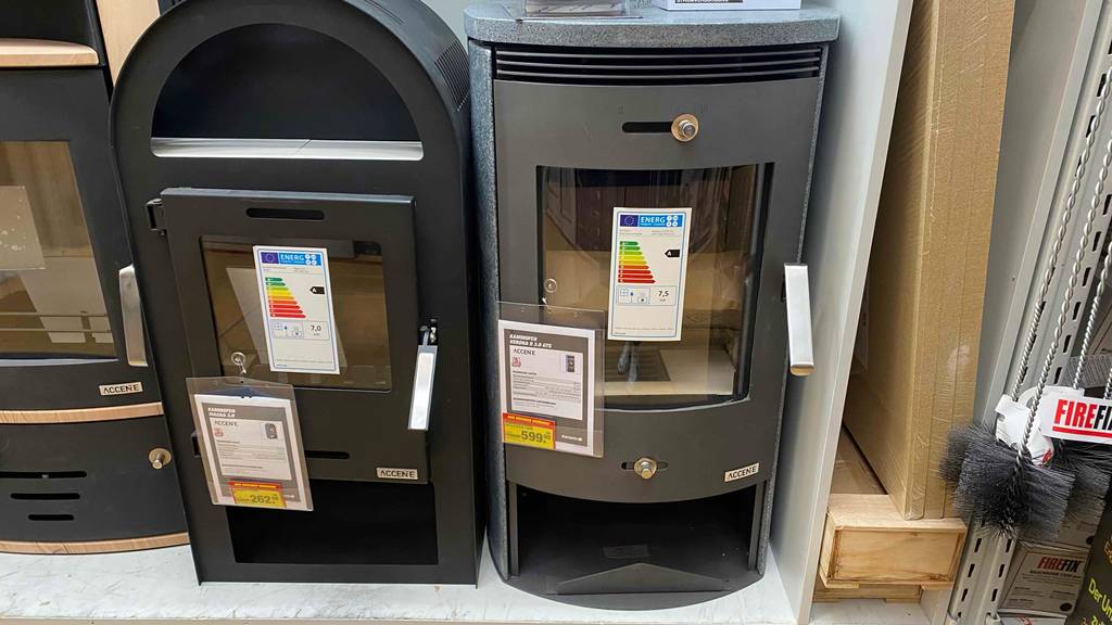 Kaminofen im Test: Ansicht von vorn auf einen anthrazit-grauen Zeitbrandofen mit Linksanschlag und großem Glasfenster neben weiteren Kaminöfen in einem Ausstellungsbereich.