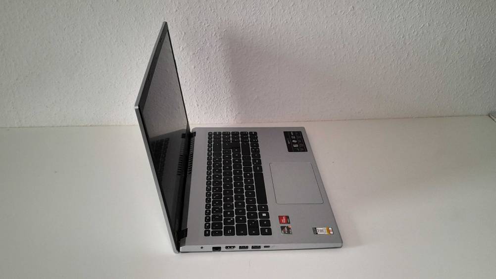 Seitenansicht von einem silberfarbenen, geöffneten 15-Zoll-Laptop im Test, das über eine schwarze Tastatur verfügt und auf einem weißen Tisch vor weißer Raufasertapete steht.