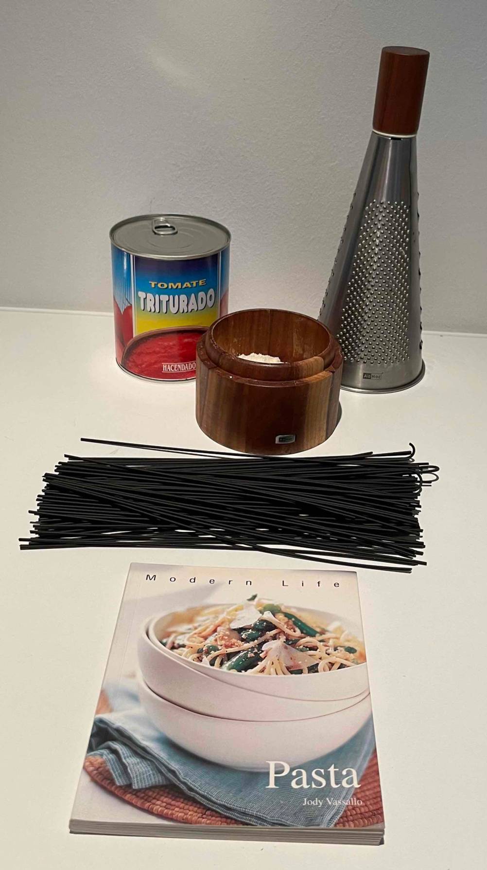 Eine Adhoc Parmesanreibe im Test befindet sich auf einer hellen Fläche zusammen mit einem Nudelkochbuch, schwarzen Spaghetti und einer Dose geschälten Tomaten.
