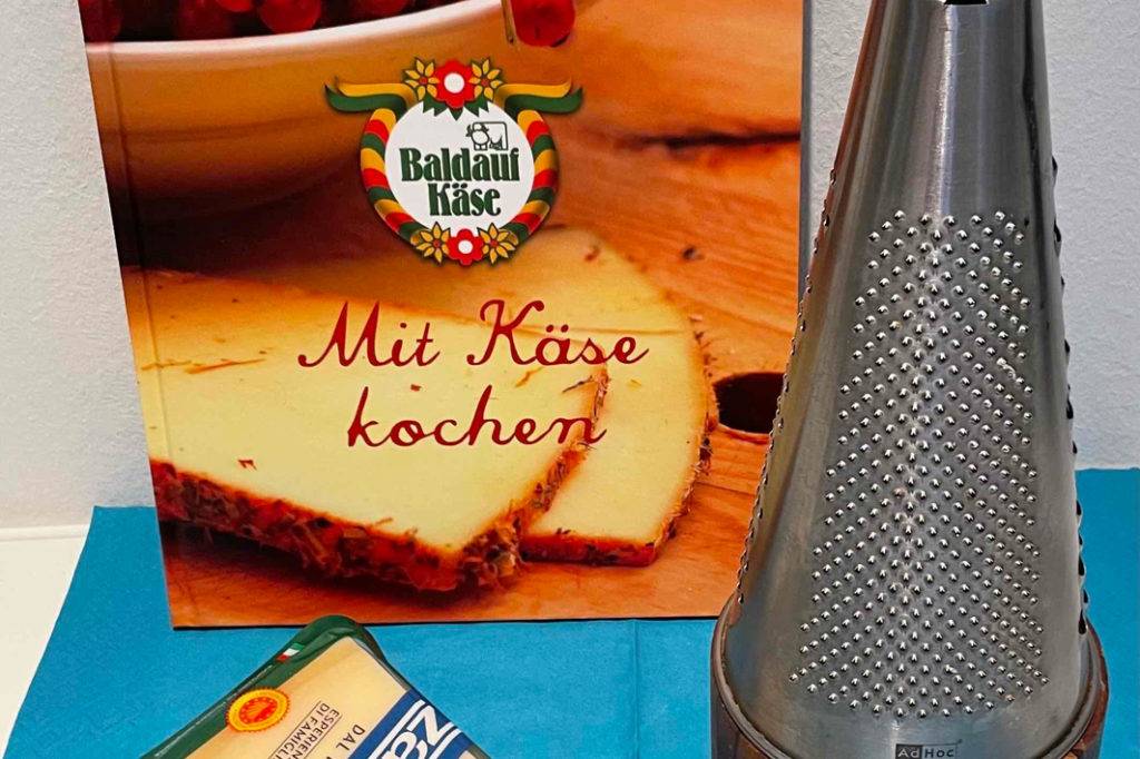 Im Test: Parmesanreibe von Adhoc steht neben Käsekochbuch und einer Packung Parmesan am Stück.