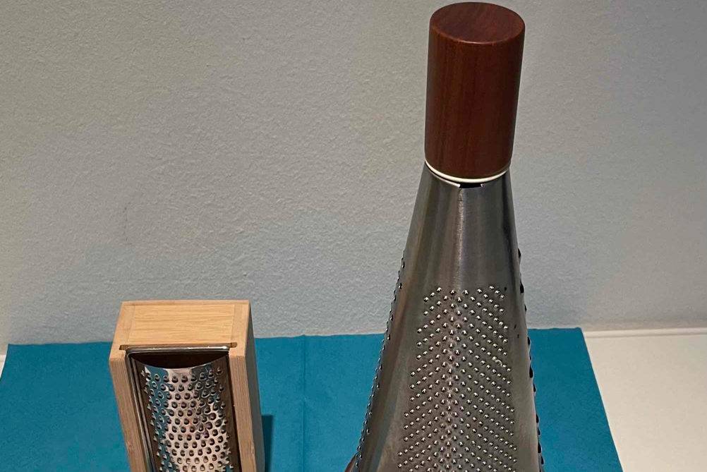 Parmesanreibe im Vergleich: mehrere verschiedene Reiben stehen mit Pasta-Kochbuch auf blauer Serviette.