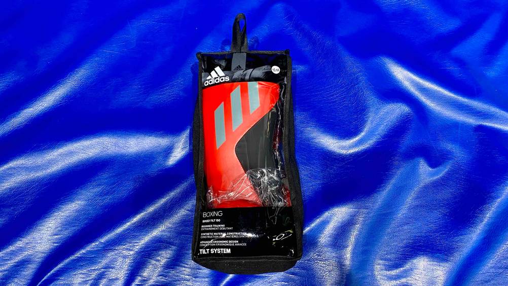 Im Test: Boxhandschuhe in schwarz-rotem Design der Marke Adidas befinden sich in einer schwarzen Verpackung inklusive Sichtfenster auf einer blauen Sportmatte.