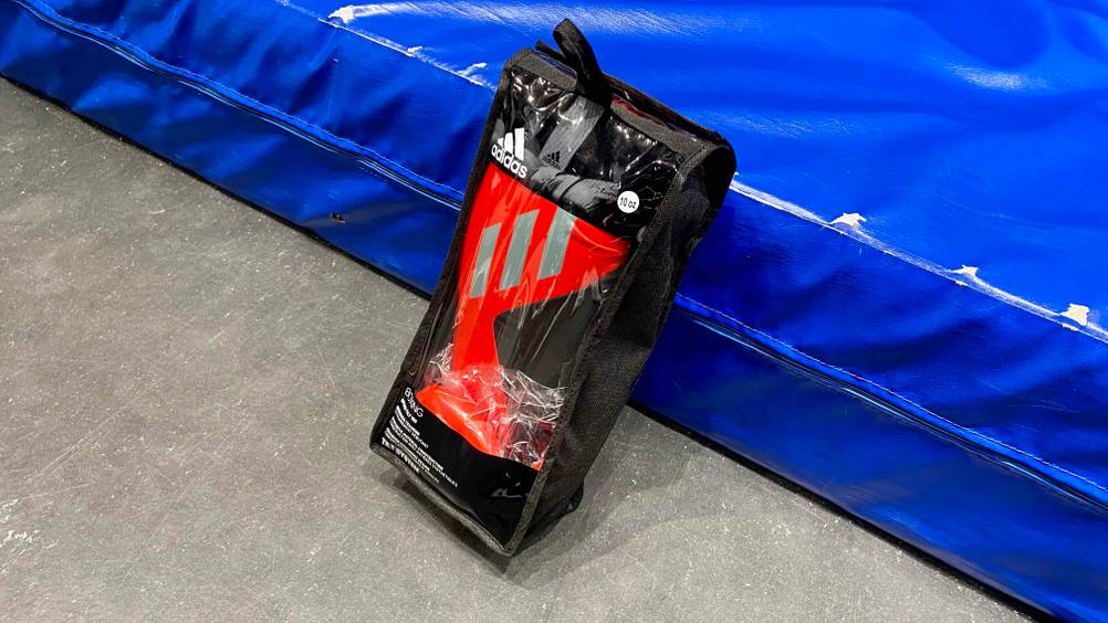 Adidas Boxhandschuhe-Test: ein schwarz-rotes Modell mit silbernen Streifen lehnt innerhalb der schwarzen Verpackung inklusive Sichtfenster am Rand einer blauen Sportmatte.