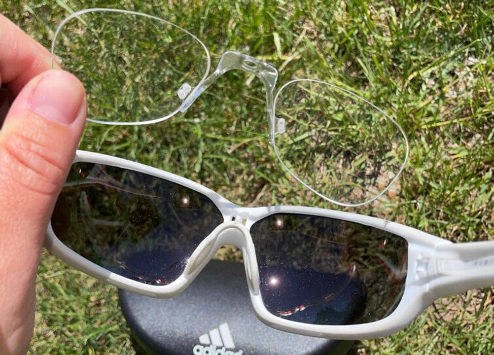 Eine Hand hält eine rahmenlose Brille über einer getesteten Fahrradbrille für Brillenträger oberhalb eines ADIDAS-Brillenetuis, das auf dem Rasen liegt.
