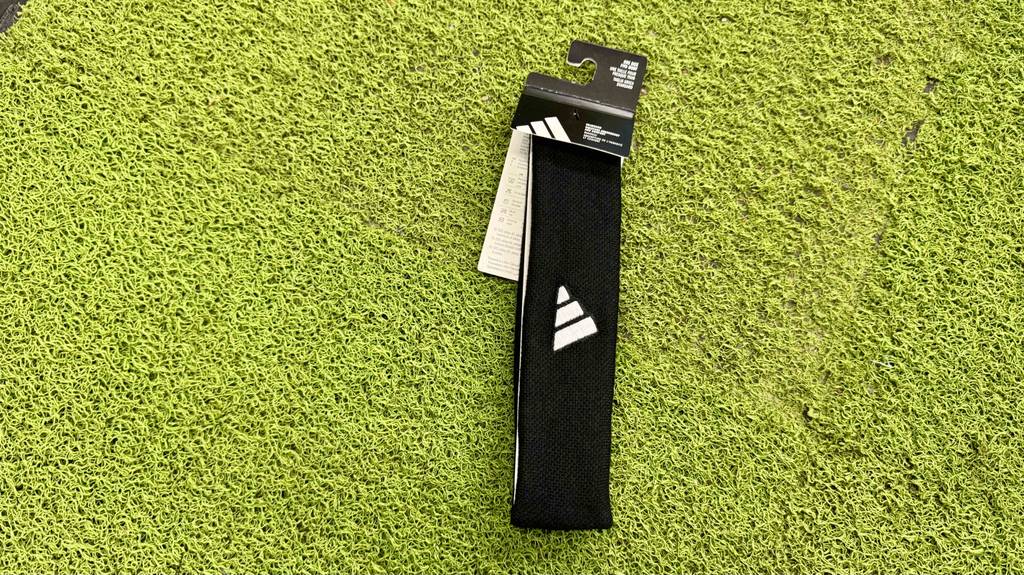 Das schwarze getestete Stirnband mit dem weißen Markenzeichen von ADIDAS liegt auf einem grünen Kunstrasen.