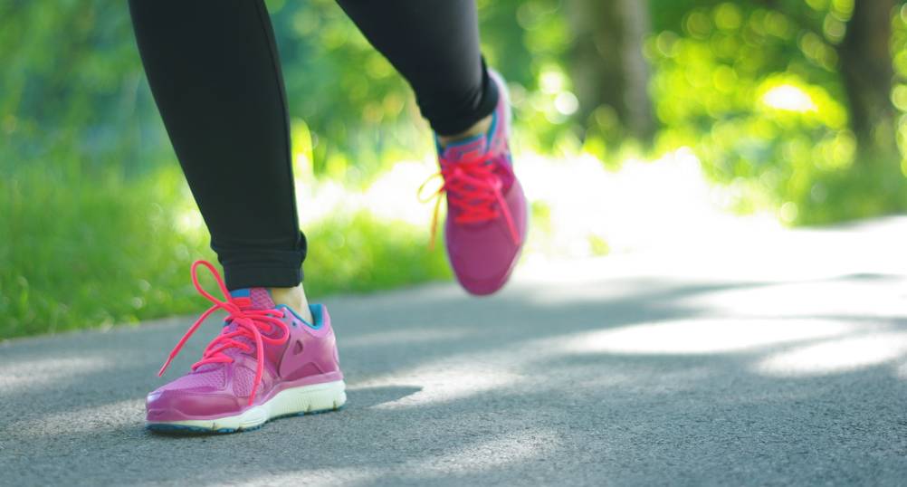 Eine Frau mit pink-orangenen Laufschuhen joggt an einem sonnigen Tag über eine Straße. Zu sehen sind nur die Unterschenkel und Füße