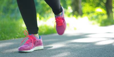 Eine Frau mit pink-orangenen Laufschuhen joggt an einem sonnigen Tag über eine Straße. Zu sehen sind nur die Unterschenkel und Füße