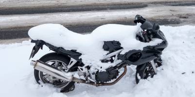 Ein Motorrad steht vor einem Holzhaus in tiefem Schnee. Auch das Motorrad ist mit einer dicken Schneeschicht bedeckt.