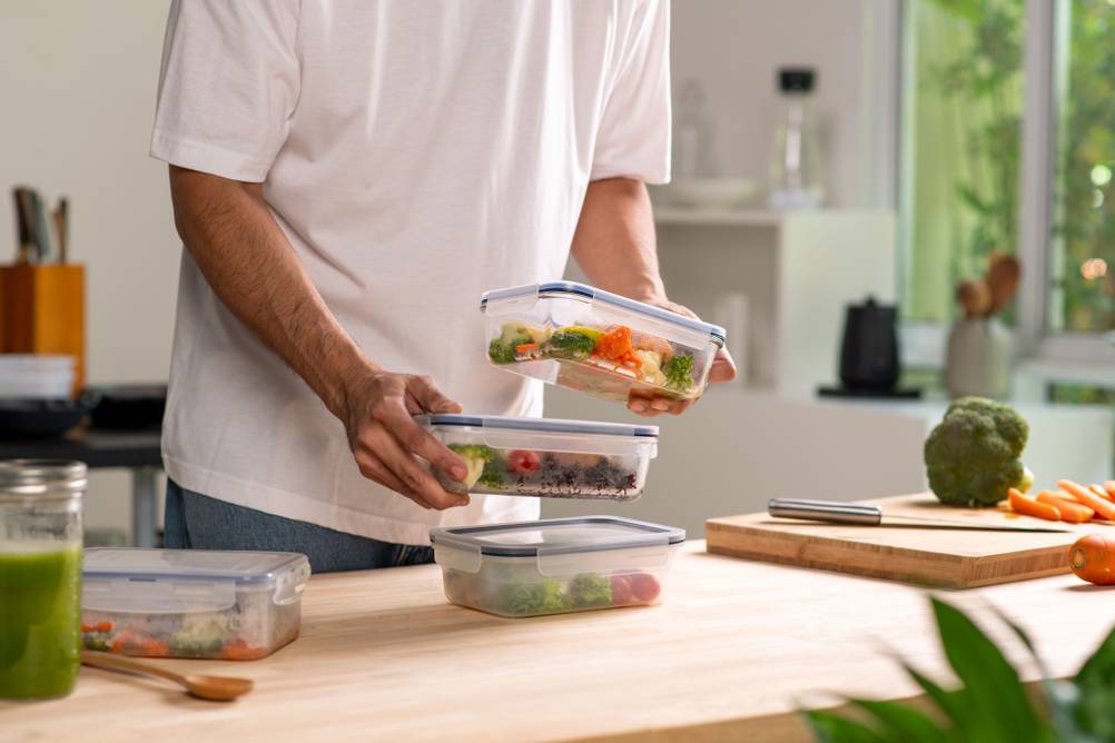 Ein Mann steht in einer Küche. Er stapelt 3 Frischhaltebehälter mit Essen übereinander auf die Kücheanrichte.