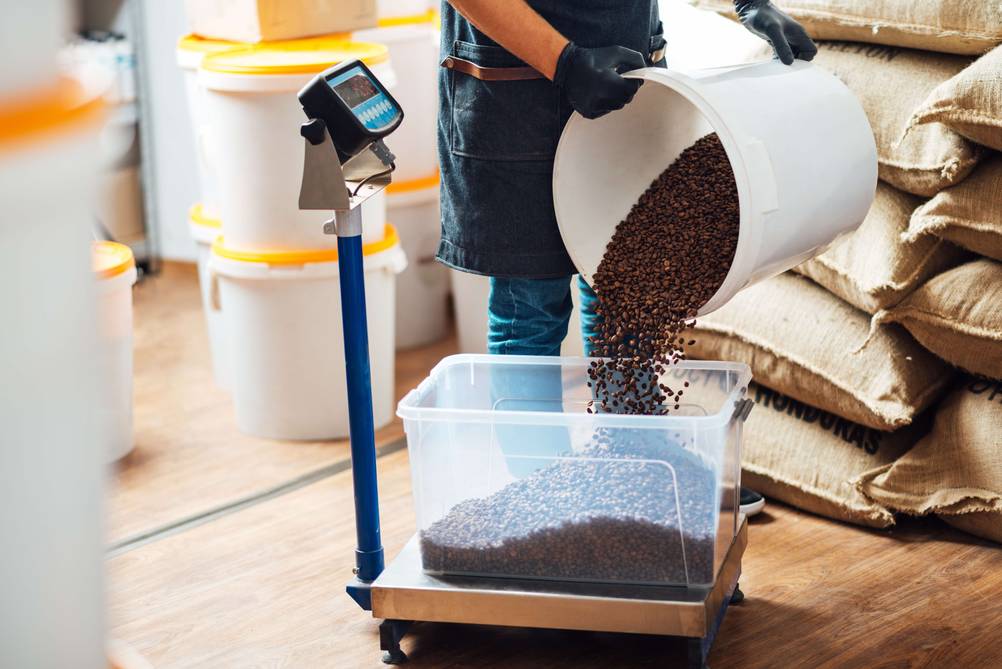 Paketwaage im Test: Mann kippt Kaffeebohnen in einen transparenten, großen Behälter der auf einer Paketwaage steht.