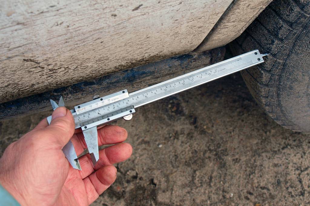 Messschieber analog im Test: Eine Männerhand hält einen analogen Messschieber mit der Tiefenmessspitze in ein Reifenprofil an einem sehr verschmutzen Kfz.