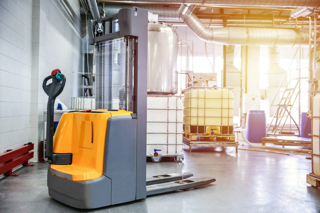 Hochhubwagen im Test: Ein Hochhubwagen steht in einer Fabrikhallte ohne Fahrer. Es stehen viele Dinge herum, wie IBC-Container, Kabel oder Schläuche. Tiefhängende Lüftungsrohre hängen von der Decke.