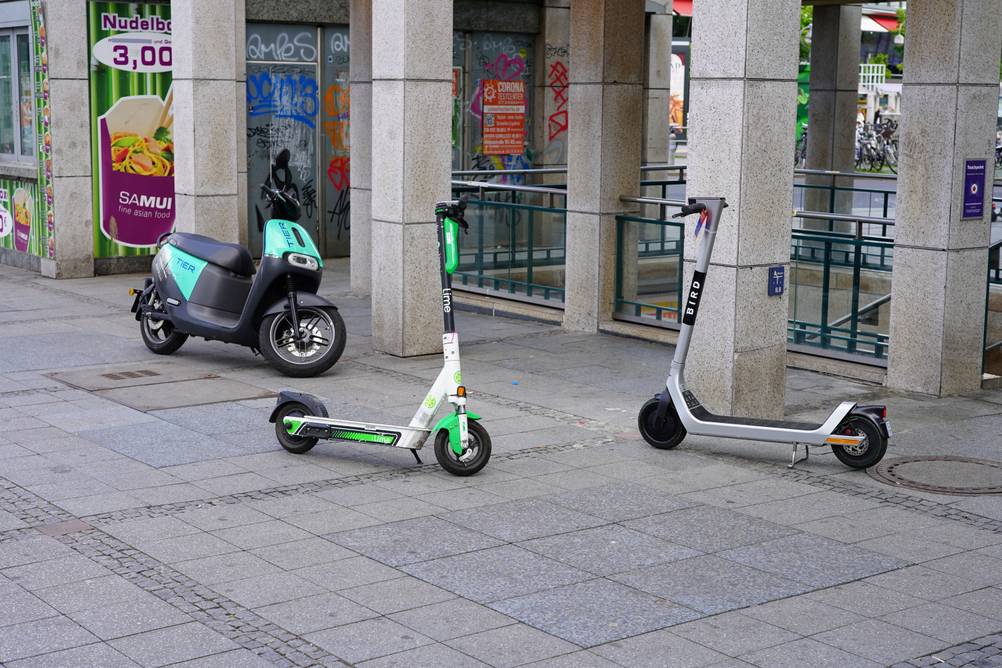 Auf einem breiten Gehweg steht ein Elektromotorrollers sowie 2 E-Scooter. Alle Fahrzeuge sind von Verleihfirmen.