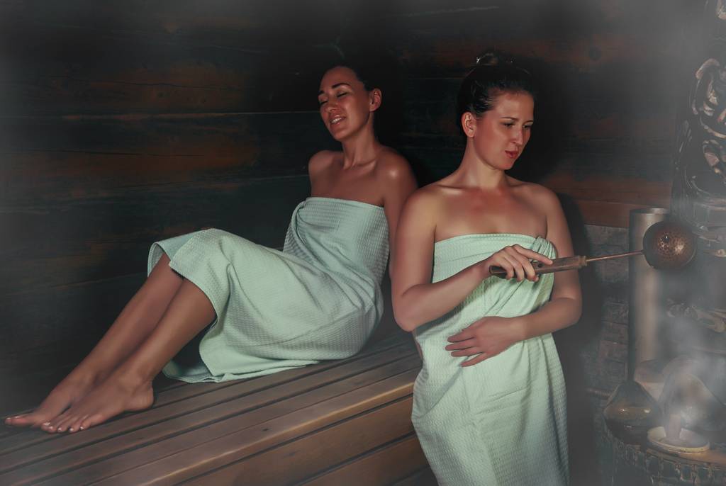 Saunaofen 6 kW im Test: Zwei Frauen in große Saunatücher gewickelt sind in der Sauna. Eine macht einen Aufguss.