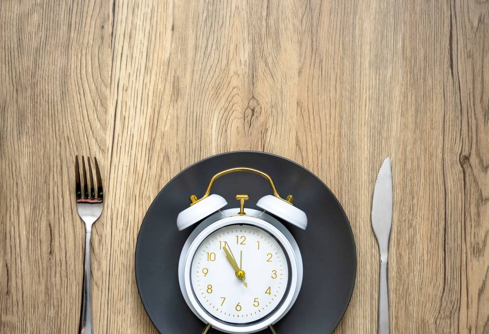 Ein Wecker liegt auf einem Teller, die Uhrzeit zeigt 5 Minuten vor 12 Uhr. Daneben liegt Besteck.