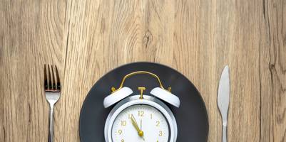 Ein Wecker liegt auf einem Teller, die Uhrzeit zeigt 5 Minuten vor 12 Uhr. Daneben liegt Besteck.