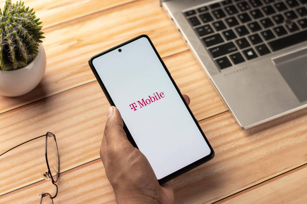 Telekom-Telefon im Test: Ein Smartphone mit dem T-Mobile-Schriftzug wird in der Hand gehalten rechts ein offenes Laptop, links ein Kaktus und eine Brille.