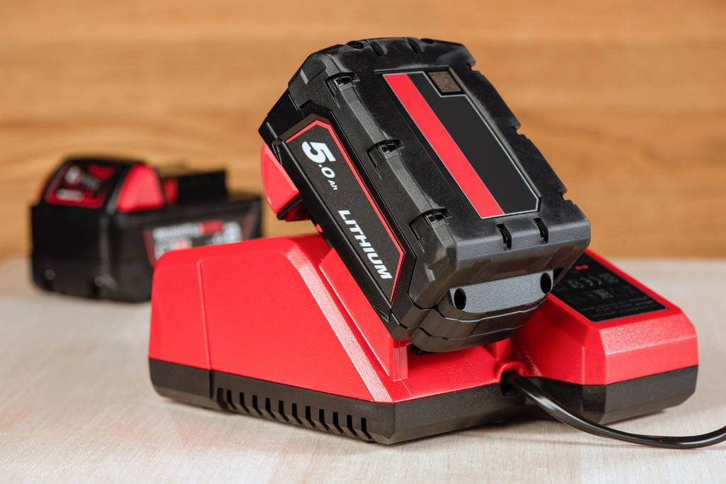 Akku-Hobel im Test: Rotes Ladegerät mit 5.0-Ah-Akku darauf und weiteren Geräten aus der Reihe im Hintergrund.