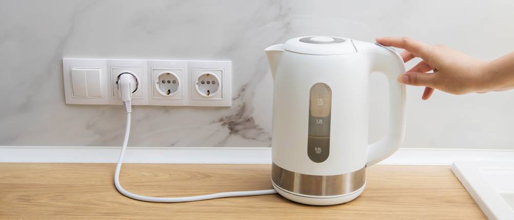 Weißer Wasserkocher im Test: Ein weißer Wasserkocher ist in eine Steckdose eingesteckt und wird von einer Hand berührt.
