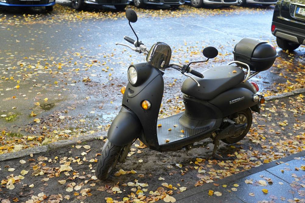 Ein E-Roller Futura parkt auf dem Bürgersteig. Die Straße ist nass. Auf dem Boden liegt gelbes Laub.