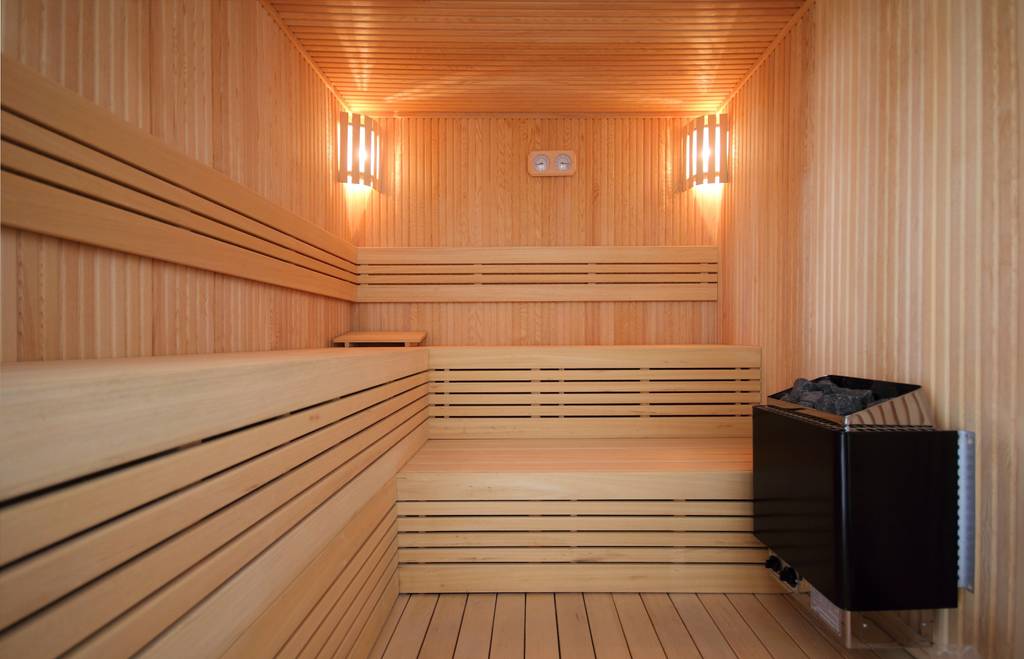 Saunaofen 6 kW im Test: Eine große, moderne Sauna sehr aufgeräumt und leer. Nur der Ofen, zwei brennende Leuchten und ein Hygro-/Thermometer sind neben den eingebauten Liegen zu sehen.