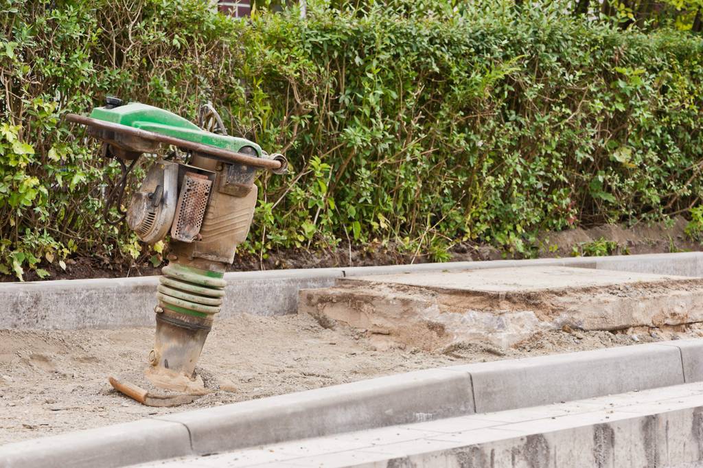 Vibrationsstampfer im Test: Ein Gerät steht auf dem Sanduntergrund eines aufgerissenen Gehwegs vor einer Hecke.