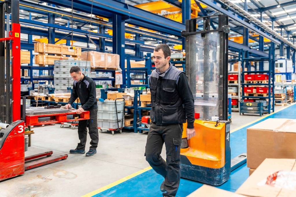Hochhubwagen im Test: Ein Mann zieht einen Hochhubwagen entlang eines blau eingezeichneten Fahrwegs in einer Fabrik.