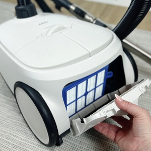 Der HEPA-Filter des AEG AB61H6SW Staubsaugers ist sichtbar, während eine Hand die hintere Klappe des Geräts öffnet. Der Staubsauger steht auf einem Teppich.