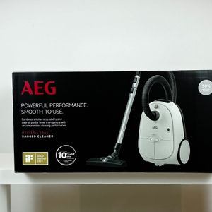 Die Verpackung des AEG AB61H6SW Staubsaugers zeigt das Produkt mit Zubehör auf schwarzem Hintergrund. Markenlogo und Features sind sichtbar.