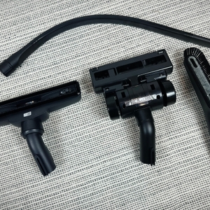 Verschiedene Zubehörteile des AEG AB61H6SW Staubsaugers liegen auf einem Teppich. Dazu gehören Bodendüsen, Fugendüse, Möbelbürste und ein flexibler Schlauch.