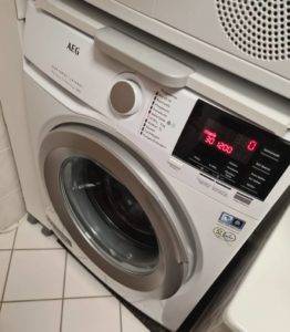 AEG Waschmaschine im Test befindet sich mit einem eingeschalteten Display unter einem Wäschetrockner im Badezimmer.